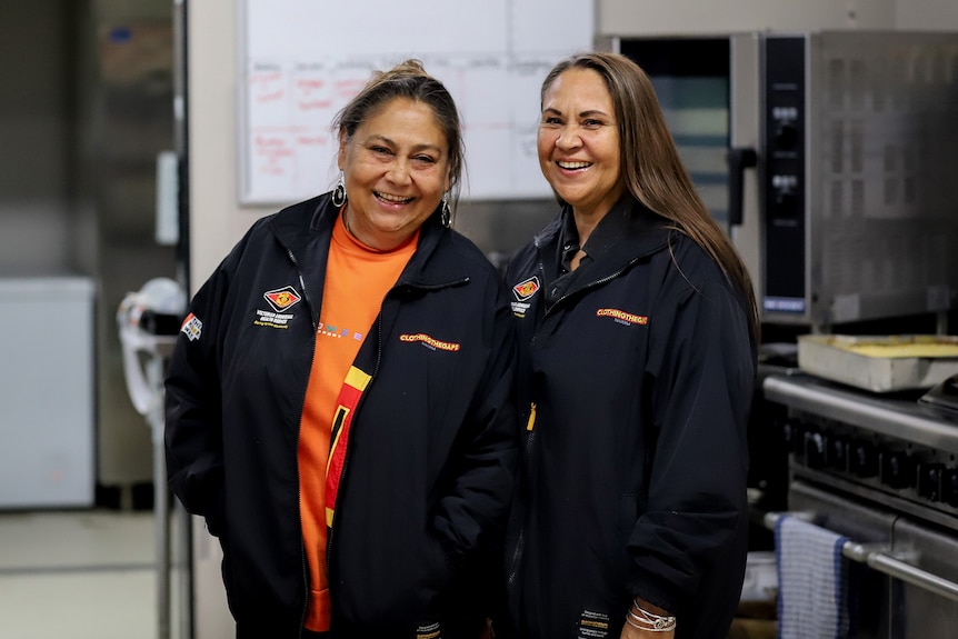 Two women smile inside a large stainless steel kitchen.