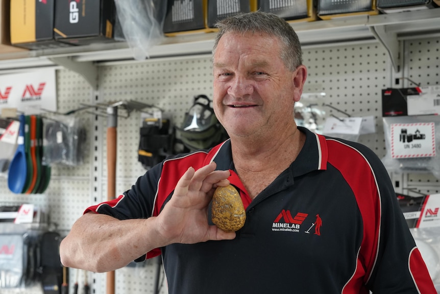 Greg Spangler standing in front of prospecting supplies and holding up the gold nugget and smiling.