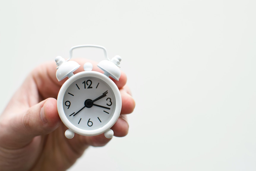 Hand holds up small white alarm clock with ticking second hand.