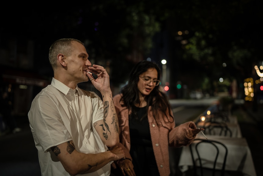 A man in a white shirt smokes a cigarette next to a woman with long hair and a dark street behind them