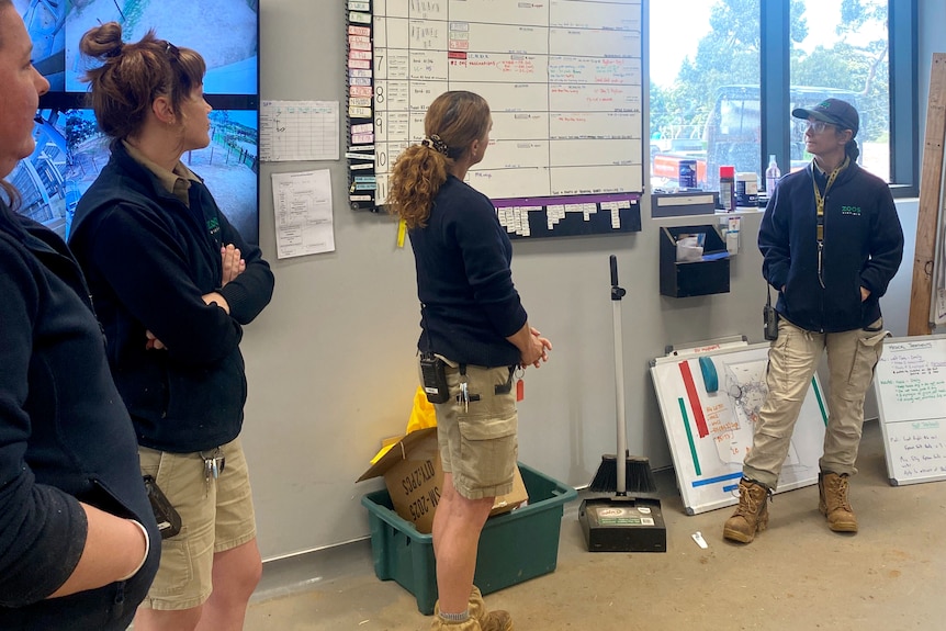 Four people in beige pants and navy jackets standing in a crib room by a whiteboard and CCTV screen.