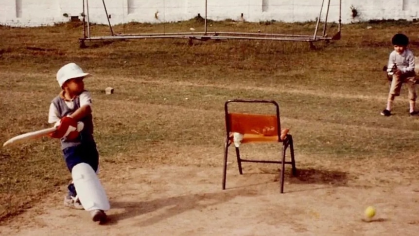 Two little boys play cricket outdoors on a dirt field. One boy bats the other fields. A chair is in the centre.