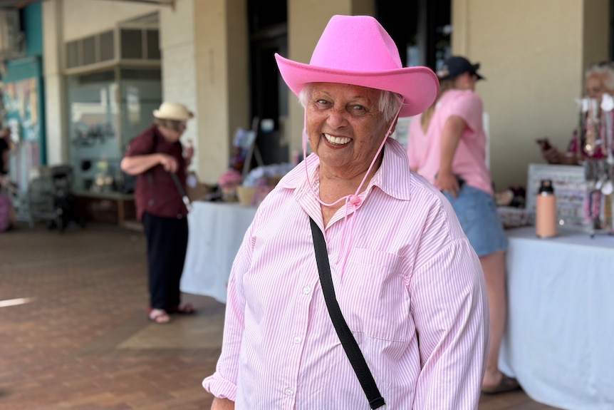 An older woman wearing a pink shirt and pink broad-brimmed hat stands in front of a store and smiles to the camera.