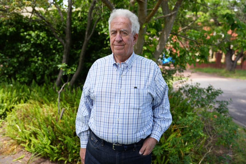 An older man in a shirt stands with hands in his pockets in a garden