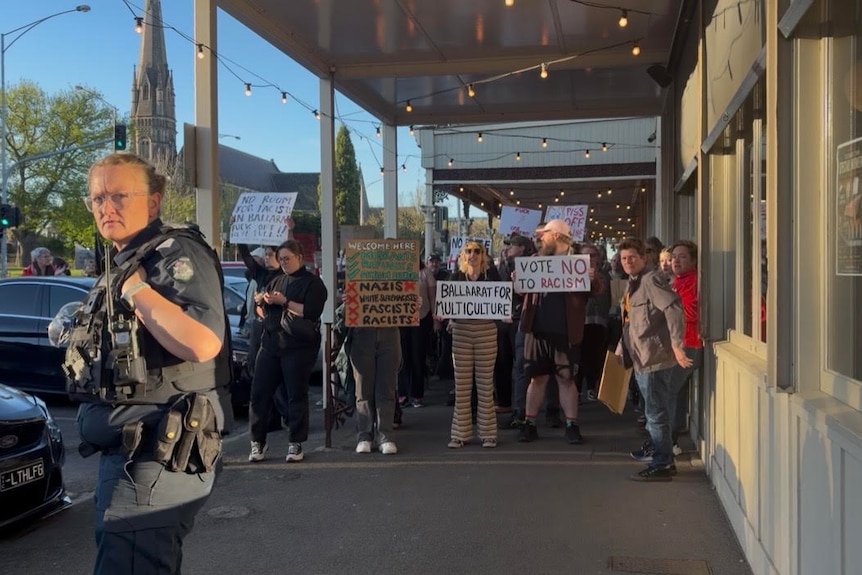 Protesters holding placards stand on a broad footpath in a country town.