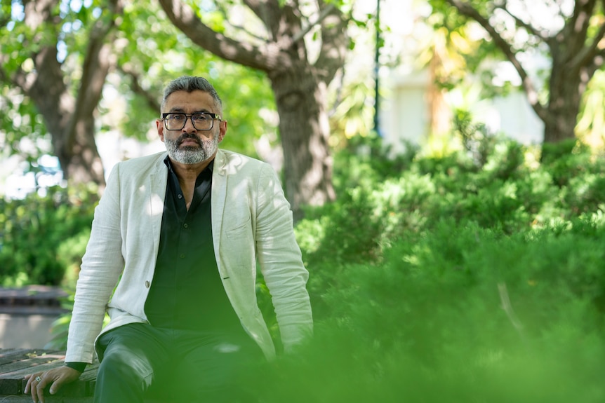 Dr Rajan Darjee sits on a bench in a leafy park of tall trees and shrubbery