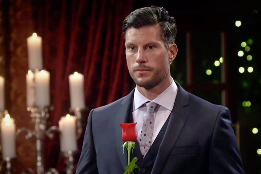Man in dark suit, with spotted tie, looking solemn and holding a single rose