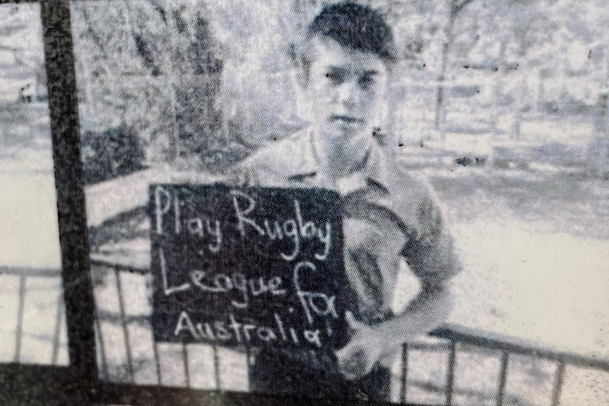 A black-and-white photo of a schoolboy holding up a slate with "Play Rugby League for Australia" chalked on it.
