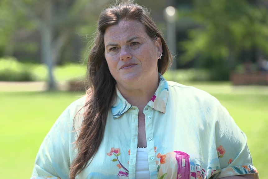 A woman with long brown hair stands outdoors looking serious.