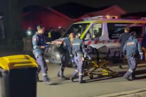 Several paramedics in overalls walk beside a stretcher to an ambulance parked on a suburban street at night.