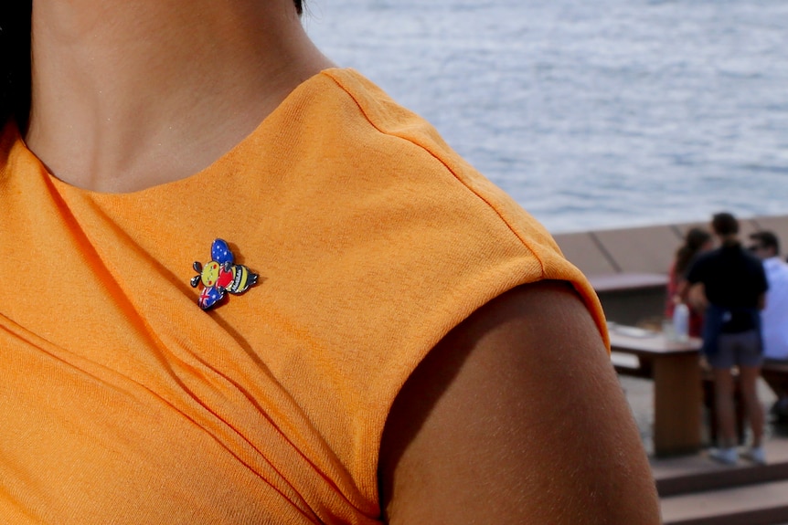 Daniella Gilboa outside the Sydney Opera House, with dark hair, an orange shirt, and a bee brooch.