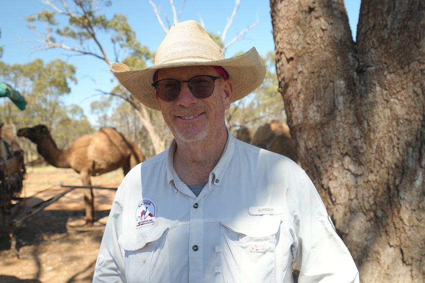 A man in a cowboy hat looks at the camera with camels in the background