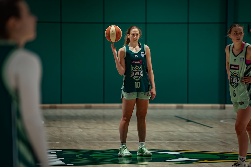 A woman in a dark green singlet in an indoor basketball court spins a basketball on her finger