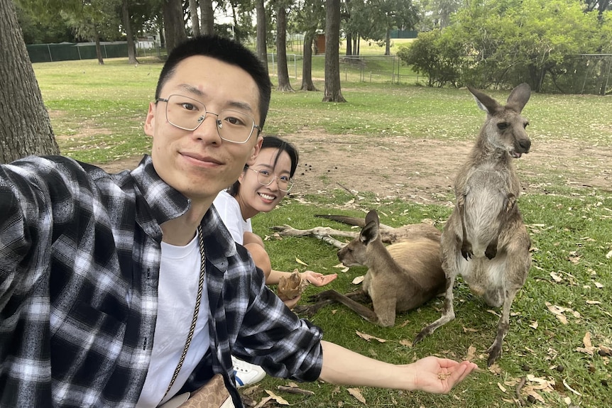 Two people take a selfie while feeding two kangaroos in a park.
