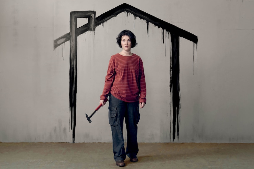 Ariadne Sgouros, early 30s, looks troubled, standing holding a hammer by her side in front of a rough painting of a house.