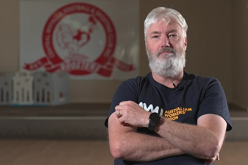 A man in a black shirt is sitting in front of an footbal club emblem which bears a red rooster image.