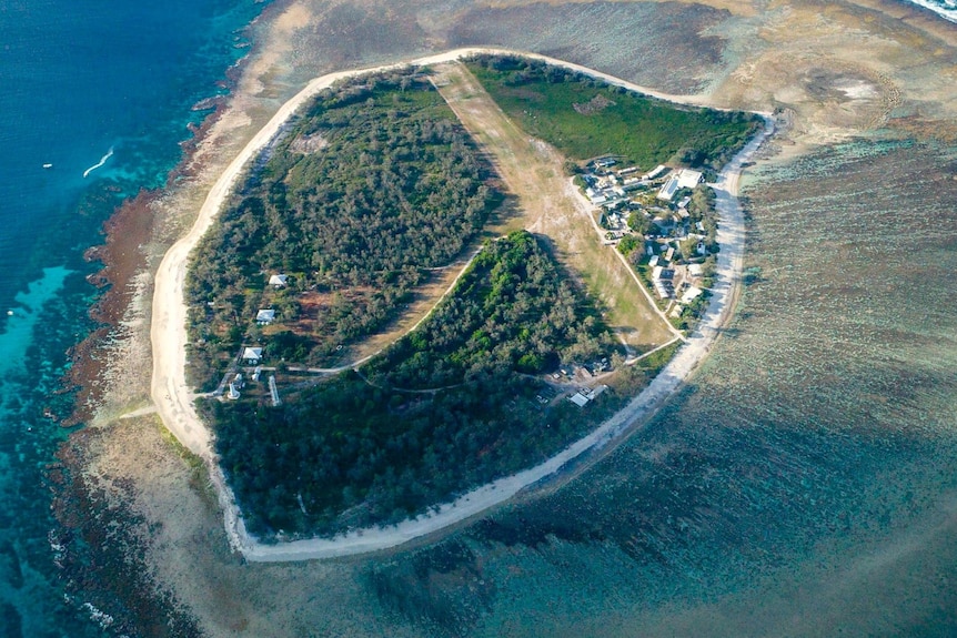 An aerial view of an island with lsuh green forests, encircled by sand and a reef.
