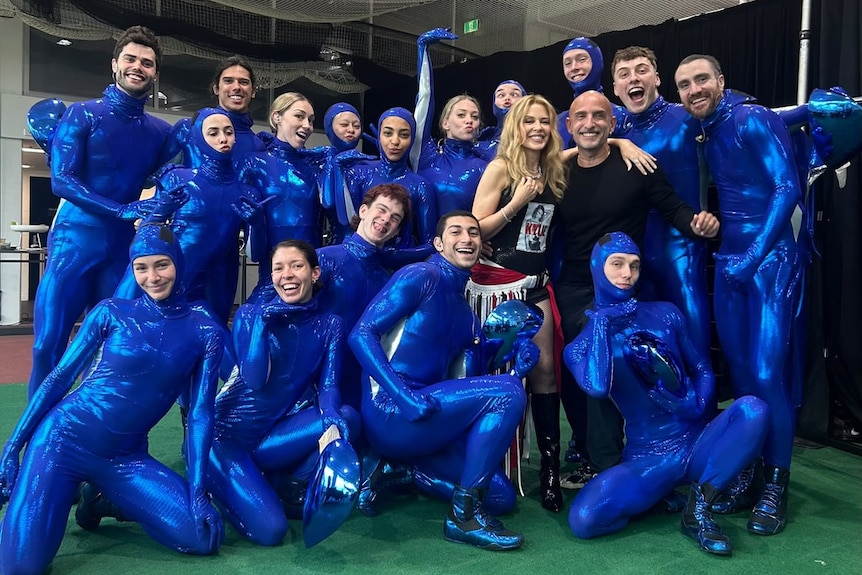 Kylie Minogue, 58, Rafael Bonachela, 54, and 16 dancers, dressed in shiny blue morph suits, pose smiling together.
