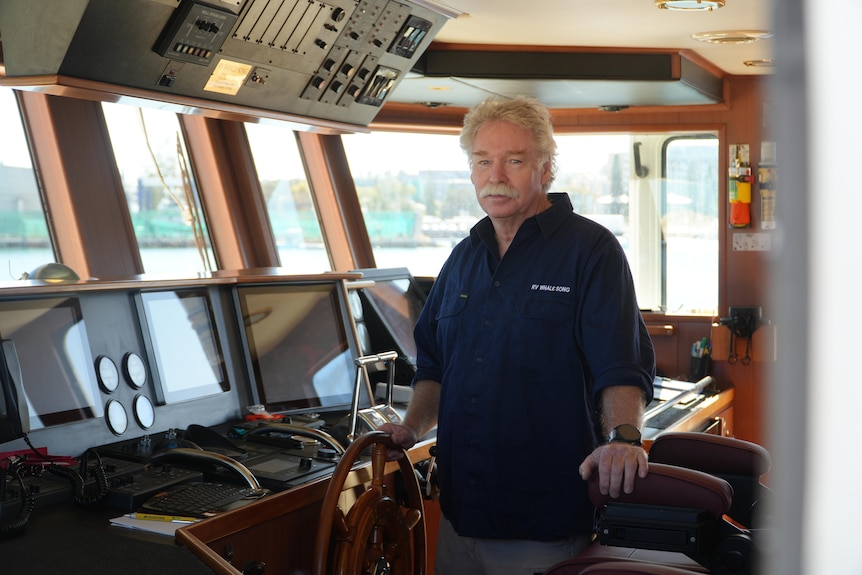 Curt wears a blue shirt as he stands in the bridge of a boat grasping the wheel