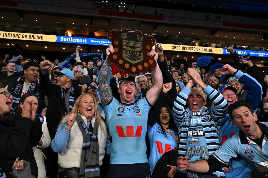 A rugby league player cheers as he lifts a championship shield above his head, surrounded by a screaming crowd.
