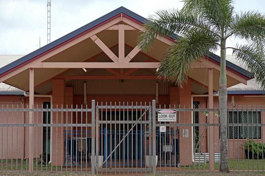 The front of a single-story brick building behind a barred fence. A palm tree stands in front. 