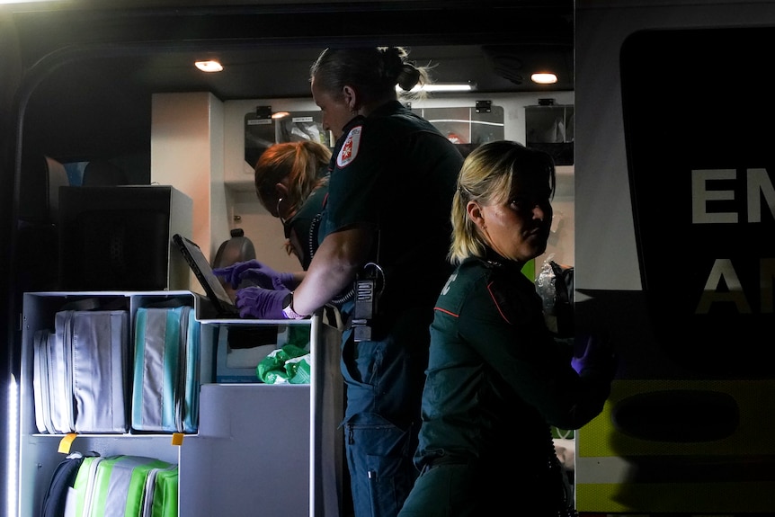 Three people in green paramedic uniforms working at the back of ambulance with its door open
