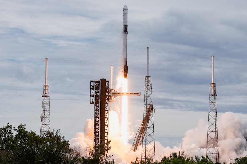A long, white space rocket firing upwards from above a blast of white-orange flame and smoke and a space launchpad tower.