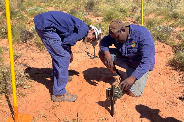 Two men dressed in dark blue ranger great setting up camera on red dirt.