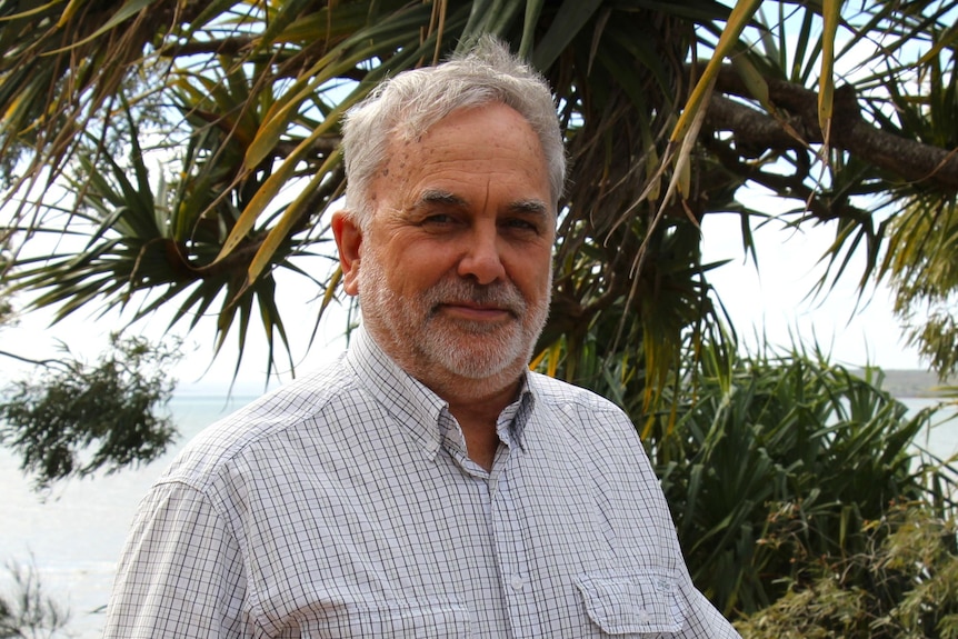 An older man with a grey beard smiles while standing in from of palm trees. 
