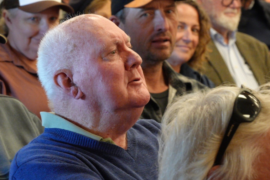 An older man sits among a crowd in a community hall.