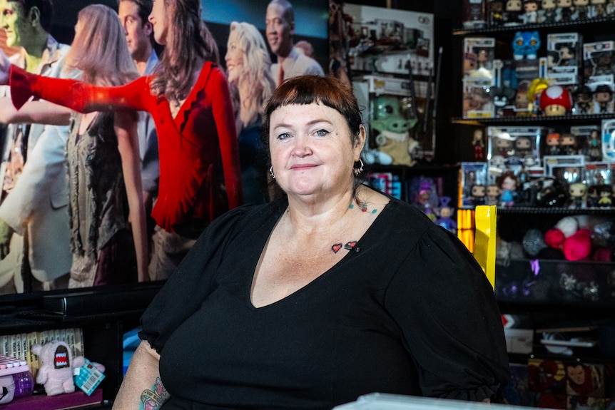 a woman in a black shirt sitting in a room with a TV over her right shoulder, and merch behind her