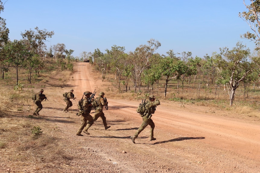 A group of people in camouflage outfits carrying weapons walking across a dusty dirt road