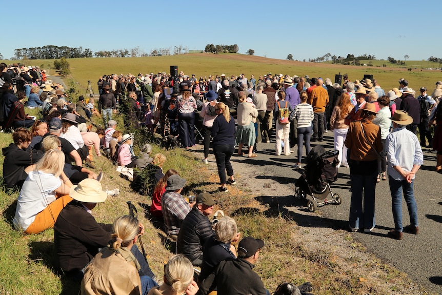 A large group of people stand and sit on a road with hills in the background.