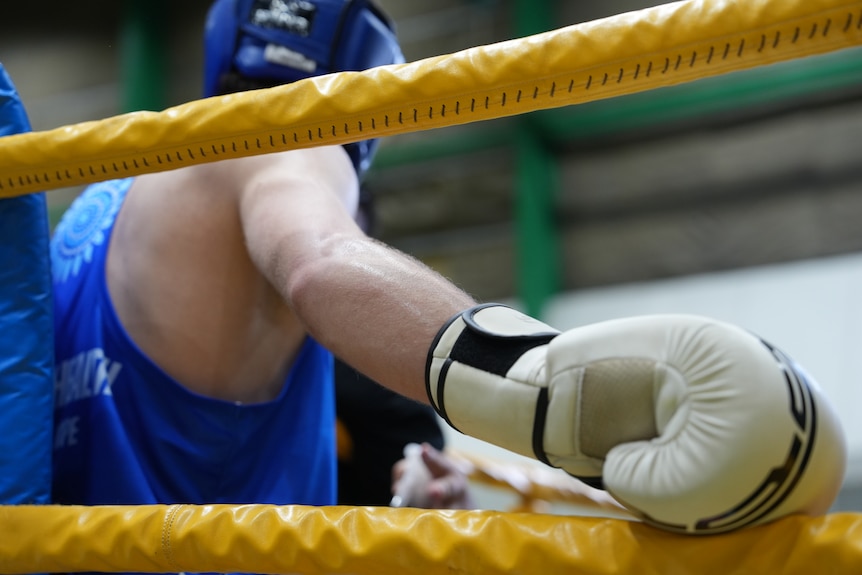 A close-up of a boxing glove as it rests on the ropes of a boxing ring, the competitor faces away from the camera.