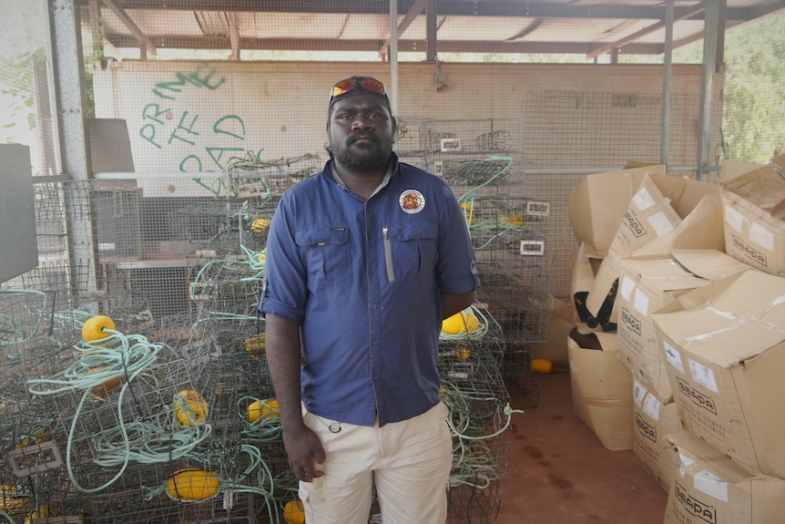 Indigenous man wearing a blue shirt and white pants standing in front of dozens of crab pots.