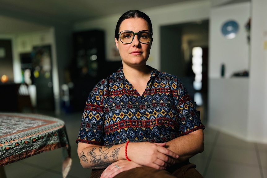 a young woman wearing a dark patterned top and glasses inside a suburban home