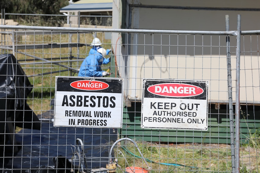 Two workers in PPE removing asbestos from side of house with asbestos signs on fence int he foreground