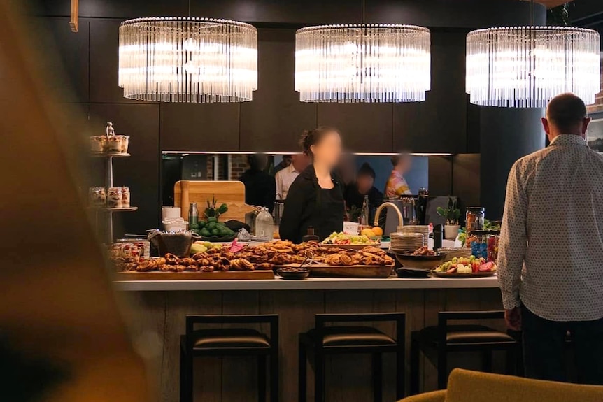 People stand around a large kitchen island covered with plates of food. Above it are three large light fixtures.