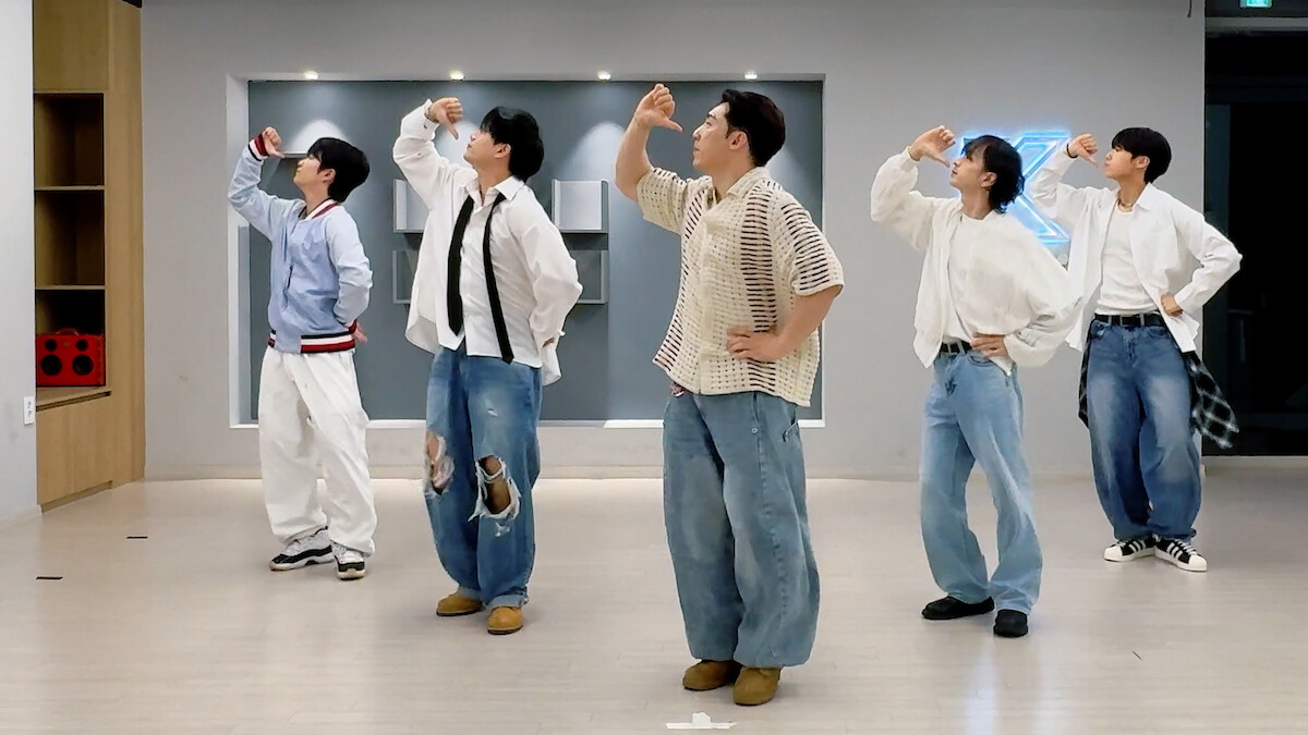 Five people in casual clothing pose in a dance studio with light wooden floors, modern decor, and a neon sign on the wall, all striking the same choreographed dance pose facing left.
