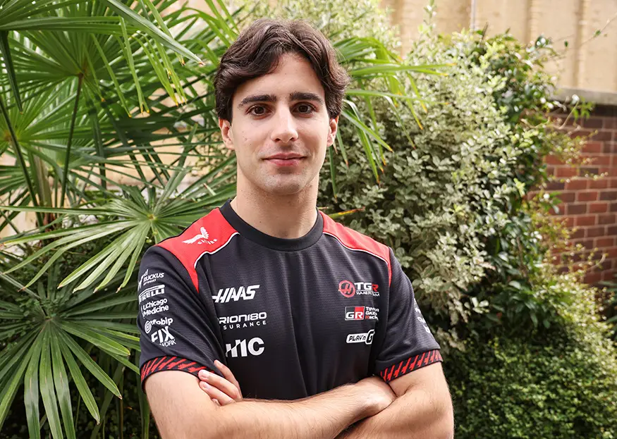 Brazilian racing driver Rafael Camara wearing a black and red TGR Haas F1 Team shirt, standing with his arms crossed in front of garden foliage.