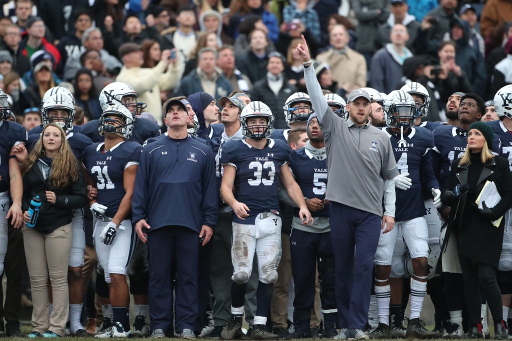 Yale football players and coaches stand on the sideline, watching intently.