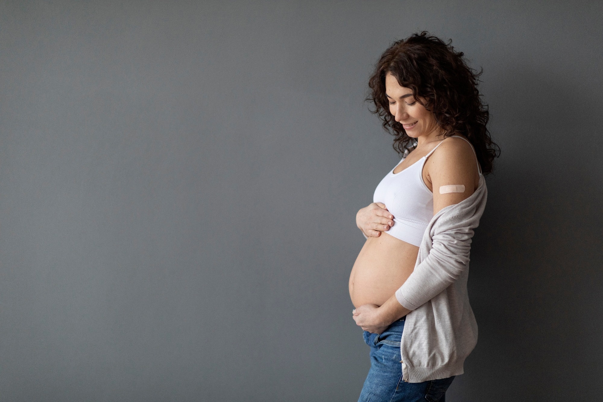 Happy Pregnant Female With Adhesive Bandage On Shoulder Embracing Her Belly While Standing Over Grey Studio background,