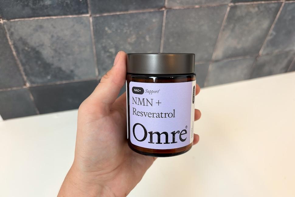 A hand holding up a glass jar of Omre NMN + Reservatrol capsules. The label is a lilac color with black lettering.