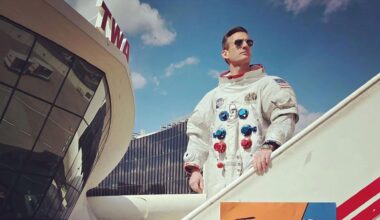 a man wearing a white spacesuit and sun glasses stands on a mobile stairway outside a 60's style airport terminal