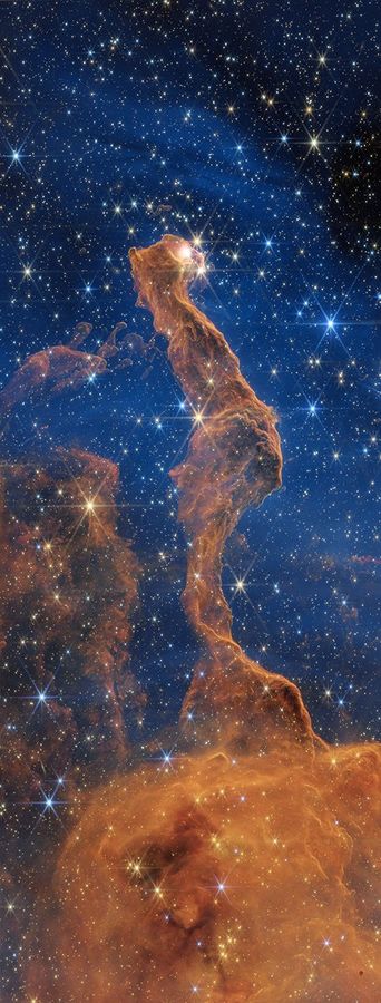 Near-infrared image of the Cat’s Head Nebula by the James Webb Space Telescope. An orange-brown filament of dust is in the center, stretching from the bottom to almost the very top. It has a narrow, irregular shape that resembles a cat sitting with its back toward the viewer and a tail extending downward. The tail ends at the lower one-third of the image and widens to a squarish dust cloud. The cat’s head is turned toward the right, revealing a yellow glowing eye. The background is mostly blue, with the top right corner revealing the black background of space. On the cat’s left are ghostly filaments of dust. Scattered throughout the view are many yellow and blue stars, some with Webb’s signature eight-point diffraction spikes, and a few faint background galaxies.