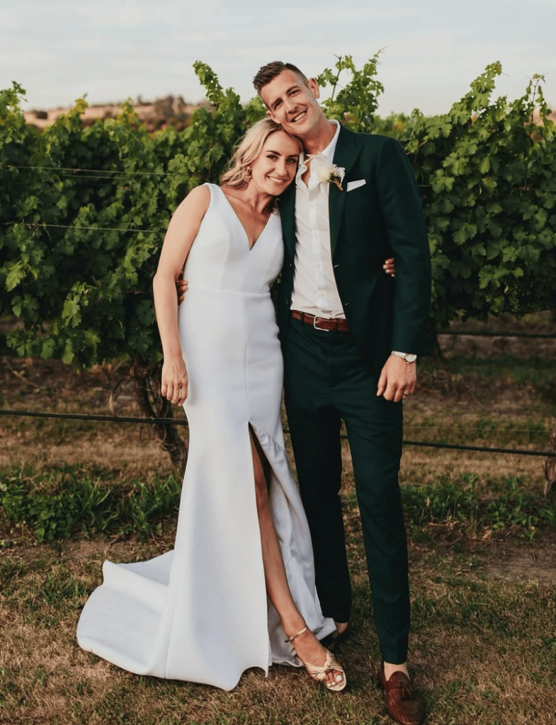 Bride and groom standing together in a vineyard, with the bride leaning her head on the groom's shoulder.