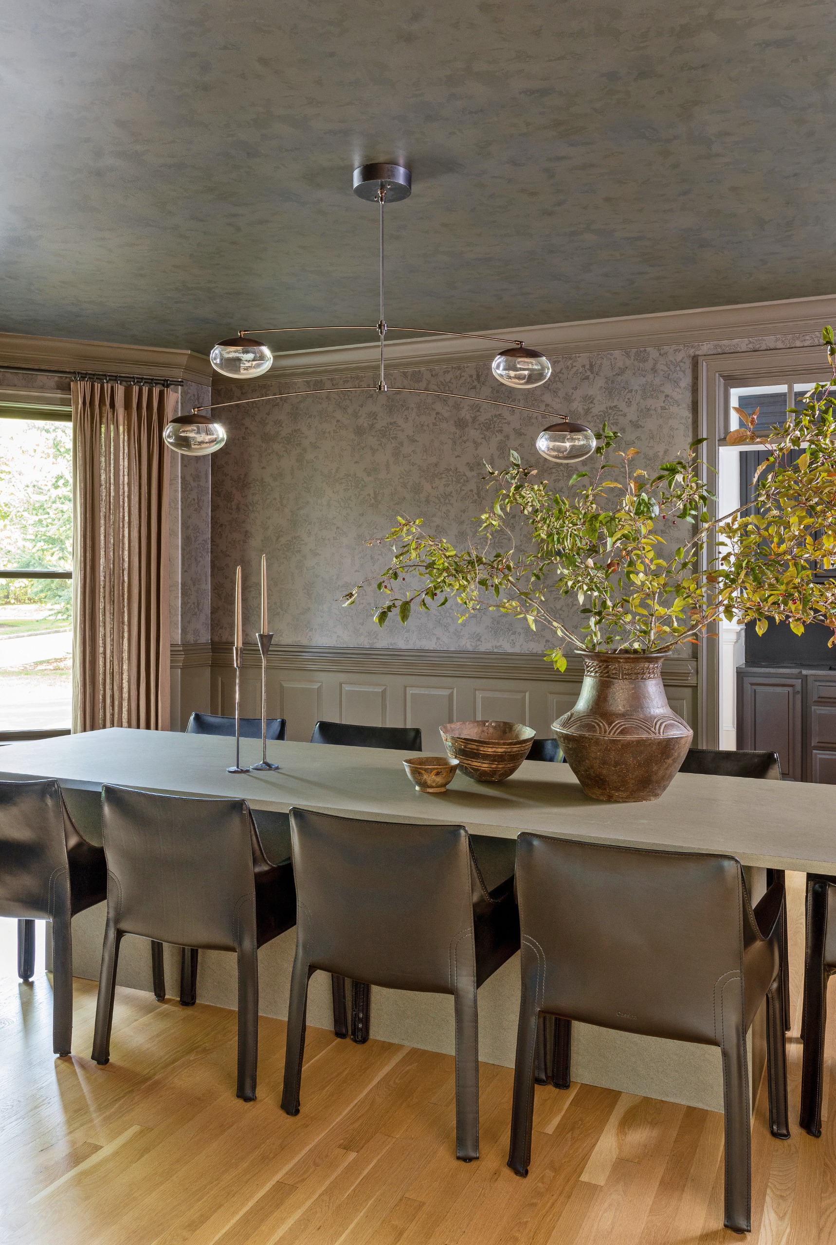 Dining room with grey patterned wallpaper and ceiling, grey concrete-look rectangular table and brown leather armchair, with brown vase on table with green foliage