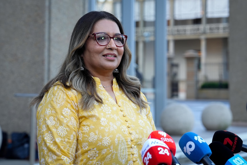 Mehreen Faruqi wearing a yellow patterned top and glasses speaking in front of microphones in front of court building.