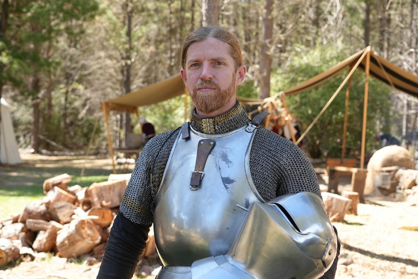 A red haired man with a beard holds a middle ages helmet and wears armour as he looks into camera 