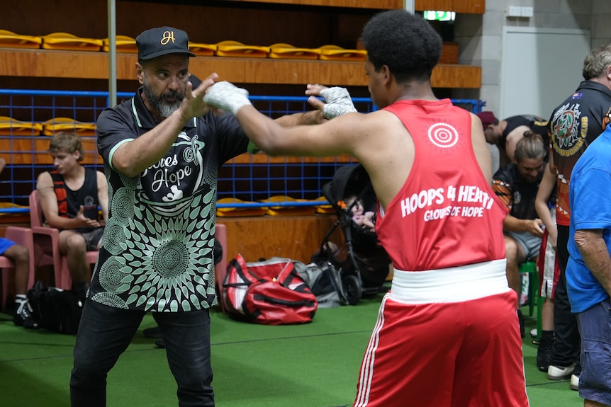 Matthew Bonson has brown skin and a beard, his hands are up in front as he blocks a young boxer's punches during warm-up.